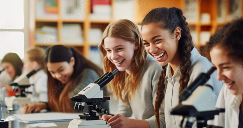 A group of students smiles and laughs while using microscopes in a science classroom. They are all seated at a long table, focusing on scientific experiments. The background shows shelves filled with books, educational materials and a science tutor. The mood is lively and cooperative. Bridging Gap Tutoring Center for Science tutoring.