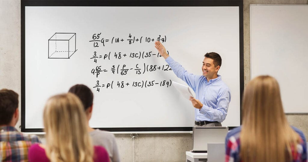 A male math tutor stands in front of a whiteboard, pointing to complex mathematical equations and a 3D cube diagram. He holds a piece of paper in his other hand. Several students are seated, facing him, with their backs visible. The classroom setting is modern and well-lit, fostering an engaging environment for math concepts. Bridging Gap Tutoring Center