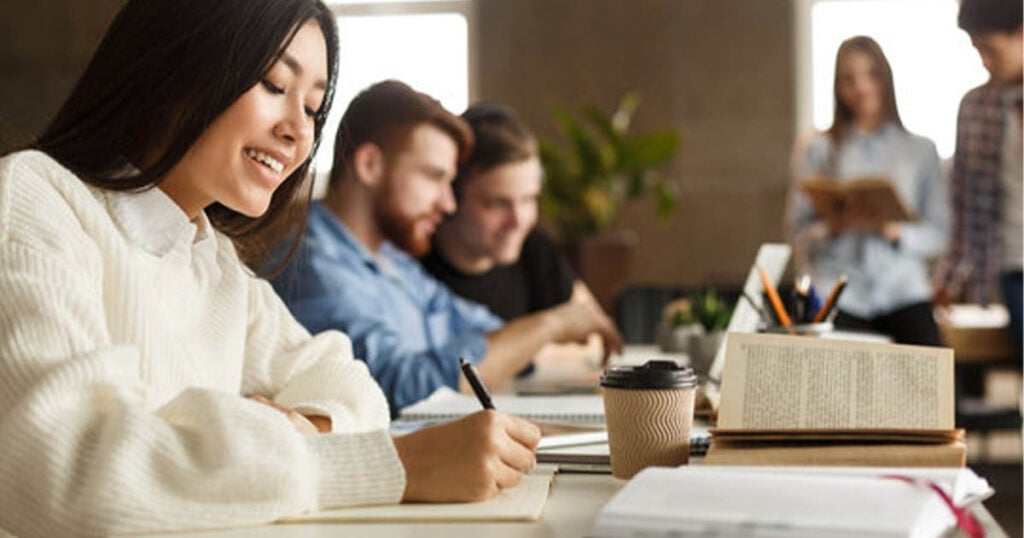 A young woman with long dark hair smiles while writing in a notebook. She is seated at a table with a laptop and a coffee cup. Behind her, two people are focused on a laptop, and two more are standing and talking, one of them holding a book. The setting appears to be a casual study environment rich in writing energy. Bridging Gap Tutoring Center
