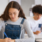 A teenage girl with long brown hair, wearing a striped shirt and denim overalls, sits at a desk writing in a classroom. Other students are seated and working in the blurred background, creating a focused and studious atmosphere. Bridging Gap Tutoring Center
