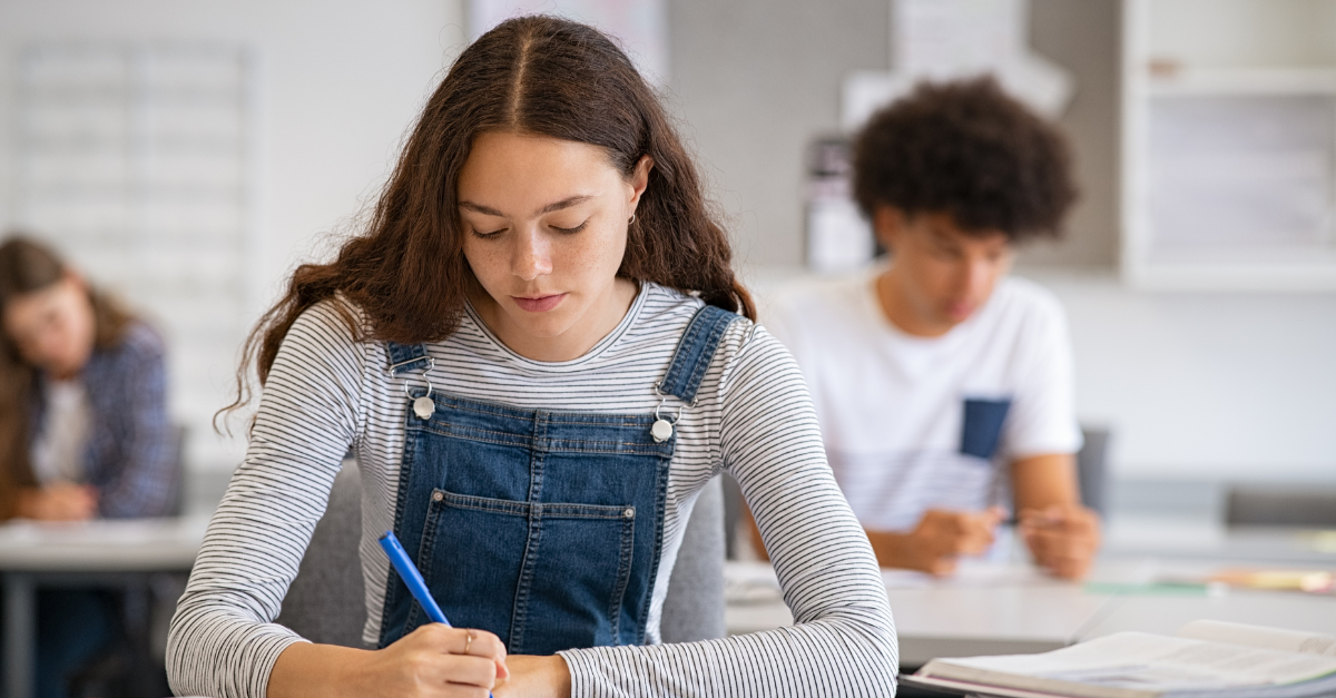 A teenage girl with long brown hair, wearing a striped shirt and denim overalls, sits at a desk writing in a classroom. Other students are seated and working in the blurred background, creating a focused and studious atmosphere. Bridging Gap Tutoring Center