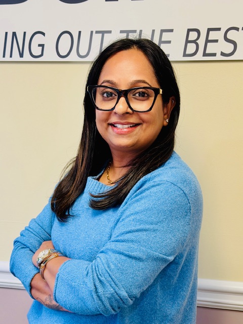 A woman with long dark hair and glasses smiles at the camera. She stands with arms crossed, wearing a light blue sweater, in front of a beige wall with a partially visible tutoring center sign behind her. Bridging Gap Tutoring Center
