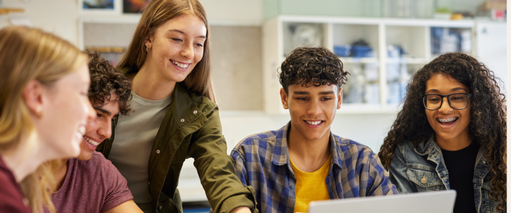 Five smiling teenagers sit together at a table in a classroom, gathered around a laptop for Georgia Milestones test prep. One girl points at the screen while the others watch, appearing engaged and happy. Shelves with supplies are visible in the background. Bridging Gap Tutoring Center