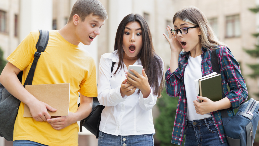 Three surprised students stand outdoors; the girl in the center, holding a phone, looks shocked. The boy on the left, in a yellow shirt, and the girl on the right, with glasses and books, both react with wide-eyed astonishment as they look at the phone. Bridging Gap Tutoring Center
